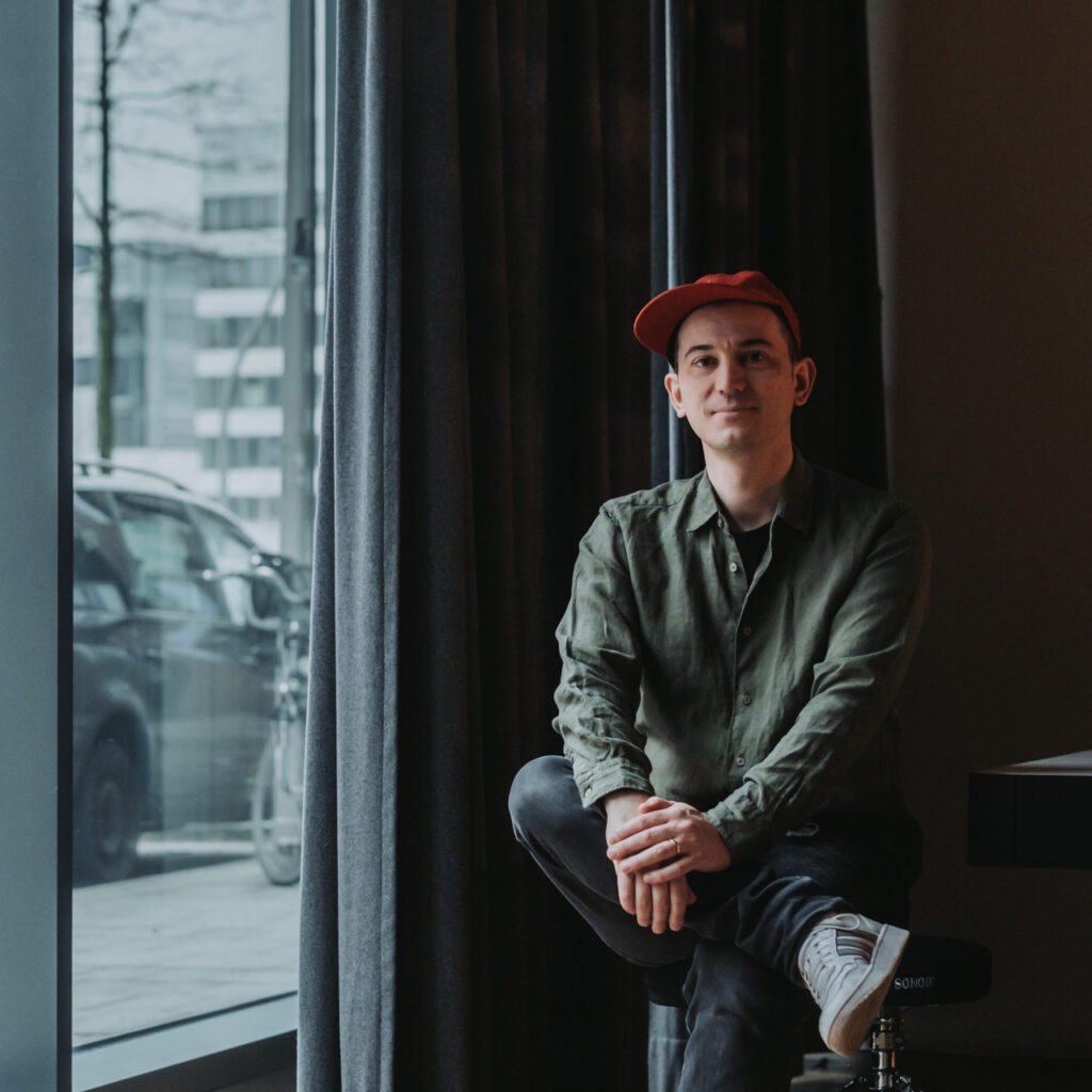 Florian-Hucht-Artdirection-Hamburg-Erfahrung-3 Florian Hucht sitz auf einem Hocker vor einem Fenster und schaut in die Kamera.